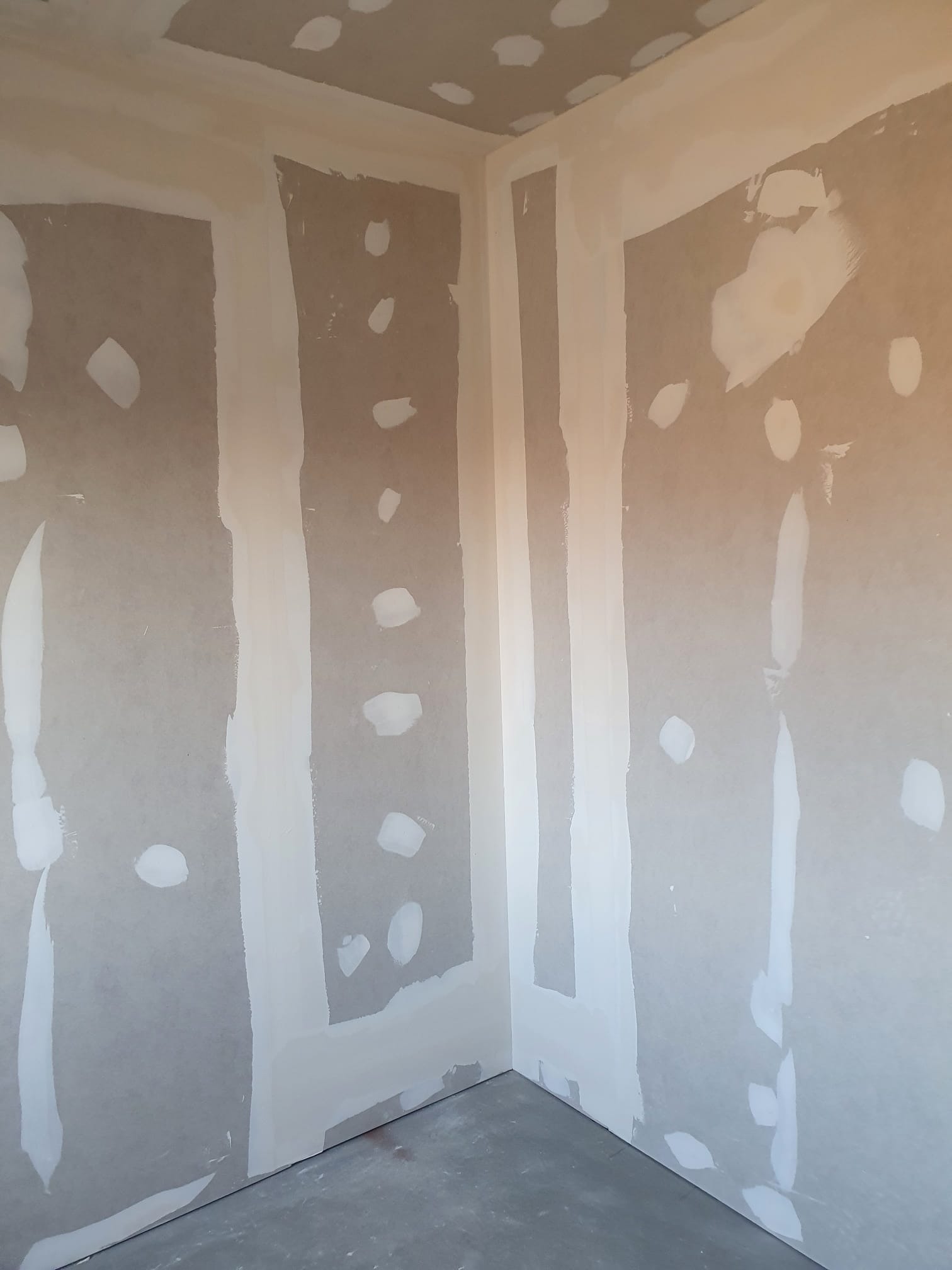 Wnętrze w trakcie wykańczania: płyty gipsowo-kartonowe z widocznymi łączeniami i miejscami po szpachlowaniu, surowy beton na podłodze.