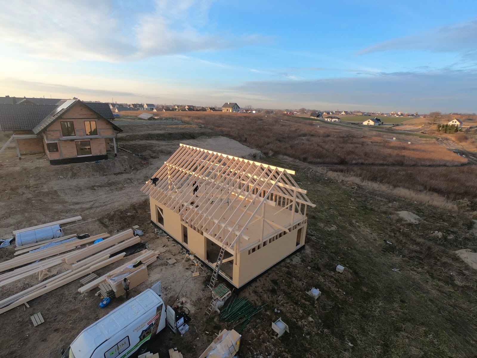 Budowa domu w technologii szkieletowej: widok z góry na konstrukcję ścian i więźby dachowej w trakcie montażu, z elementami rusztowania i stosem drewna na placu budowy, w tle zabudowa jednorodzinna...