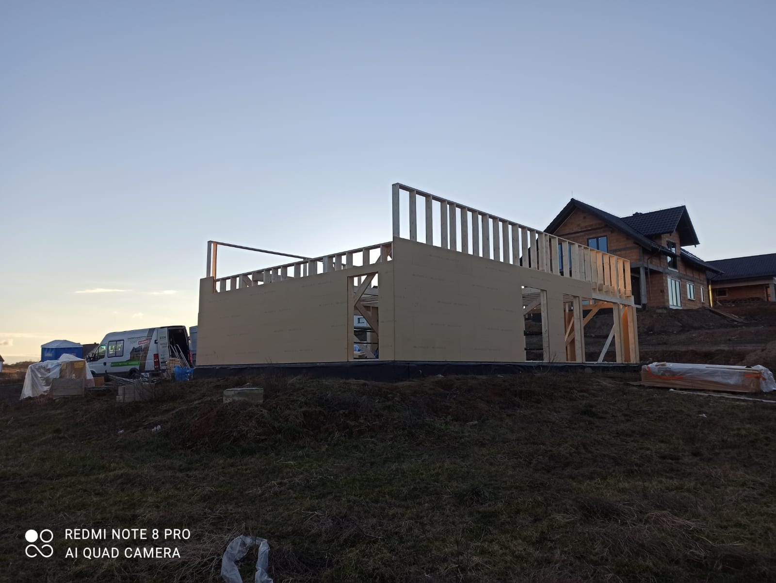 Wznoszenie konstrukcji domu z drewnianych paneli, widoczny szkielet budynku na tle zachodzącego słońca i częściowo wykończonego domu obok.