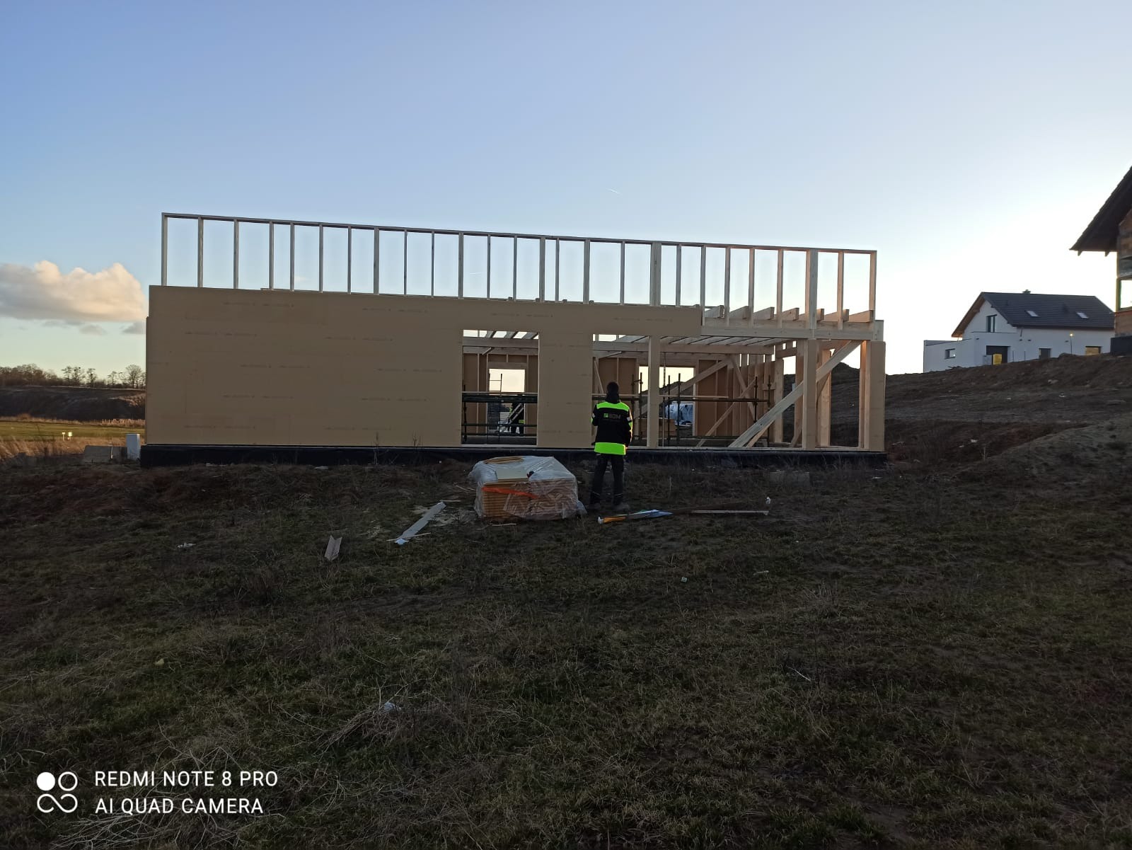 Wznoszenie konstrukcji szkieletowej domu jednorodzinnego na fundamencie, widoczny pracownik w odblaskowej kamizelce i stos desek. W tle krajobraz z zabudową mieszkaniową.