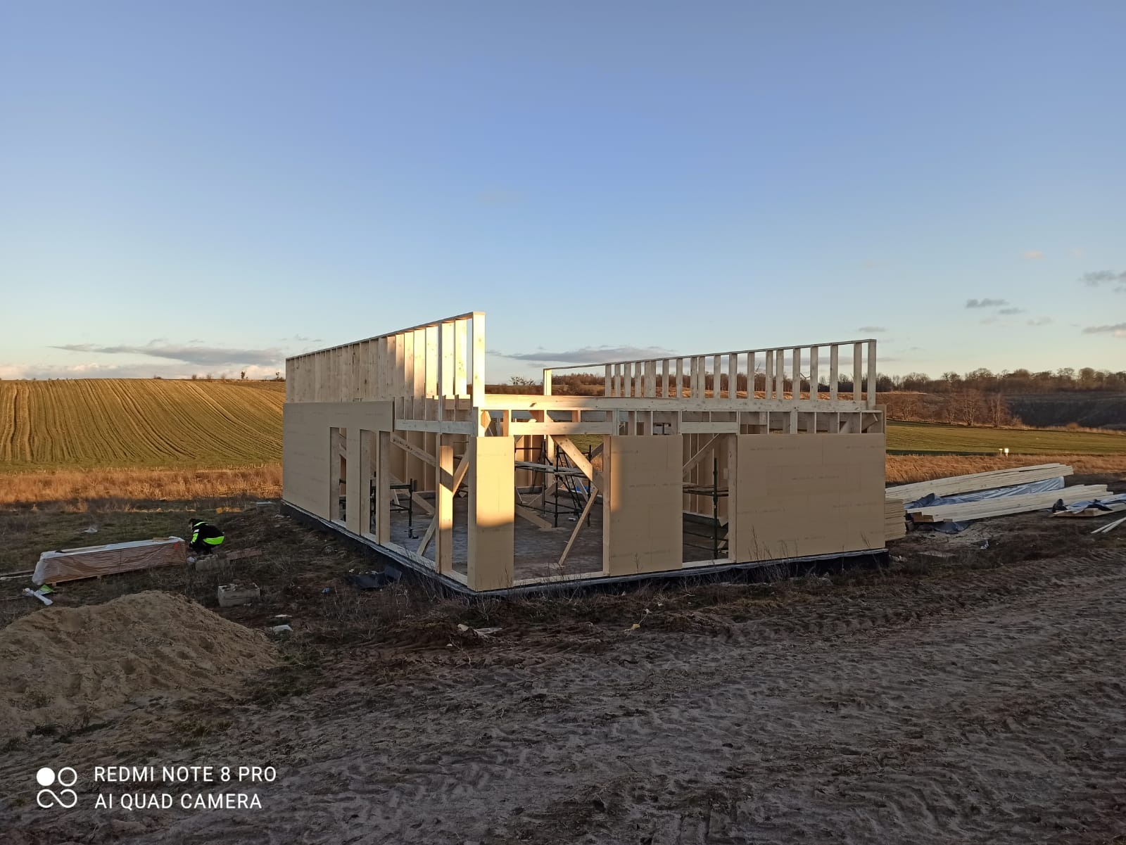 Wznoszenie szkieletu drewnianego domu w Sierakowicach, widoczne elementy konstrukcyjne ścian i stropu drugiego piętra na tle pola i błękitnego nieba.