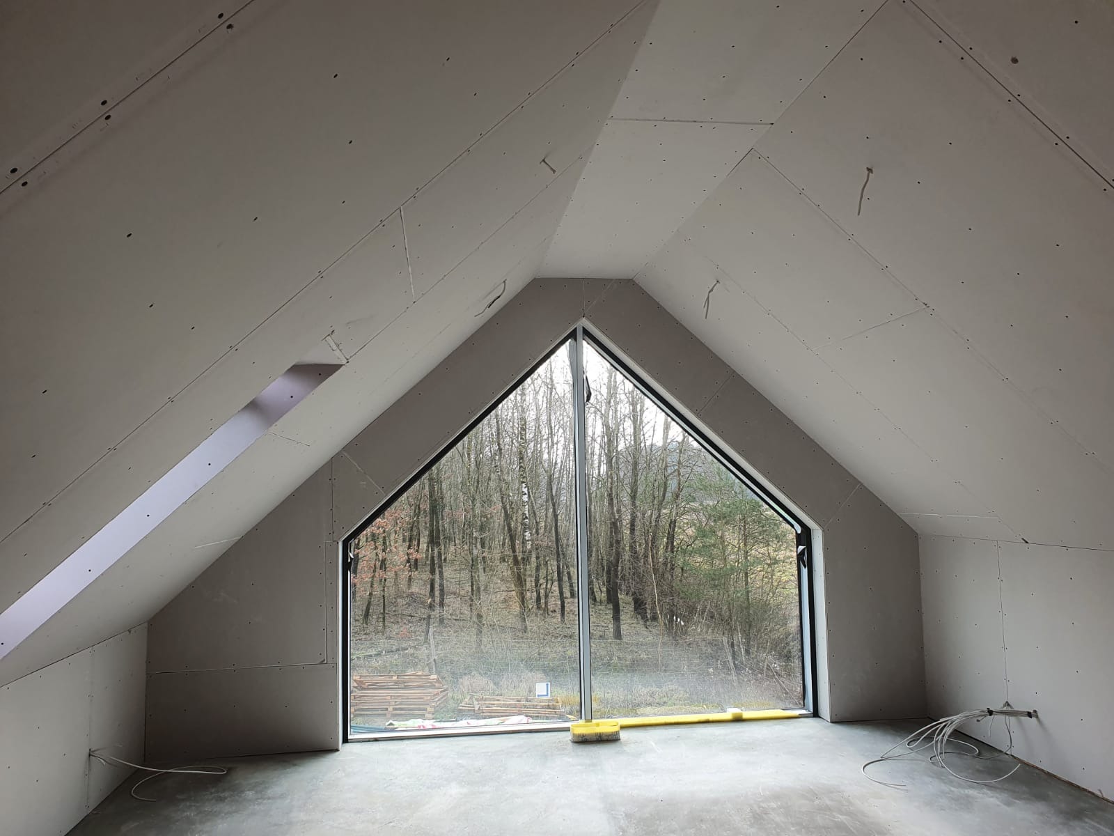 Poddasze w trakcie wykańczania płytami gipsowo-kartonowymi, widoczne okno szczytowe z widokiem na las i stertę drewna na zewnątrz.