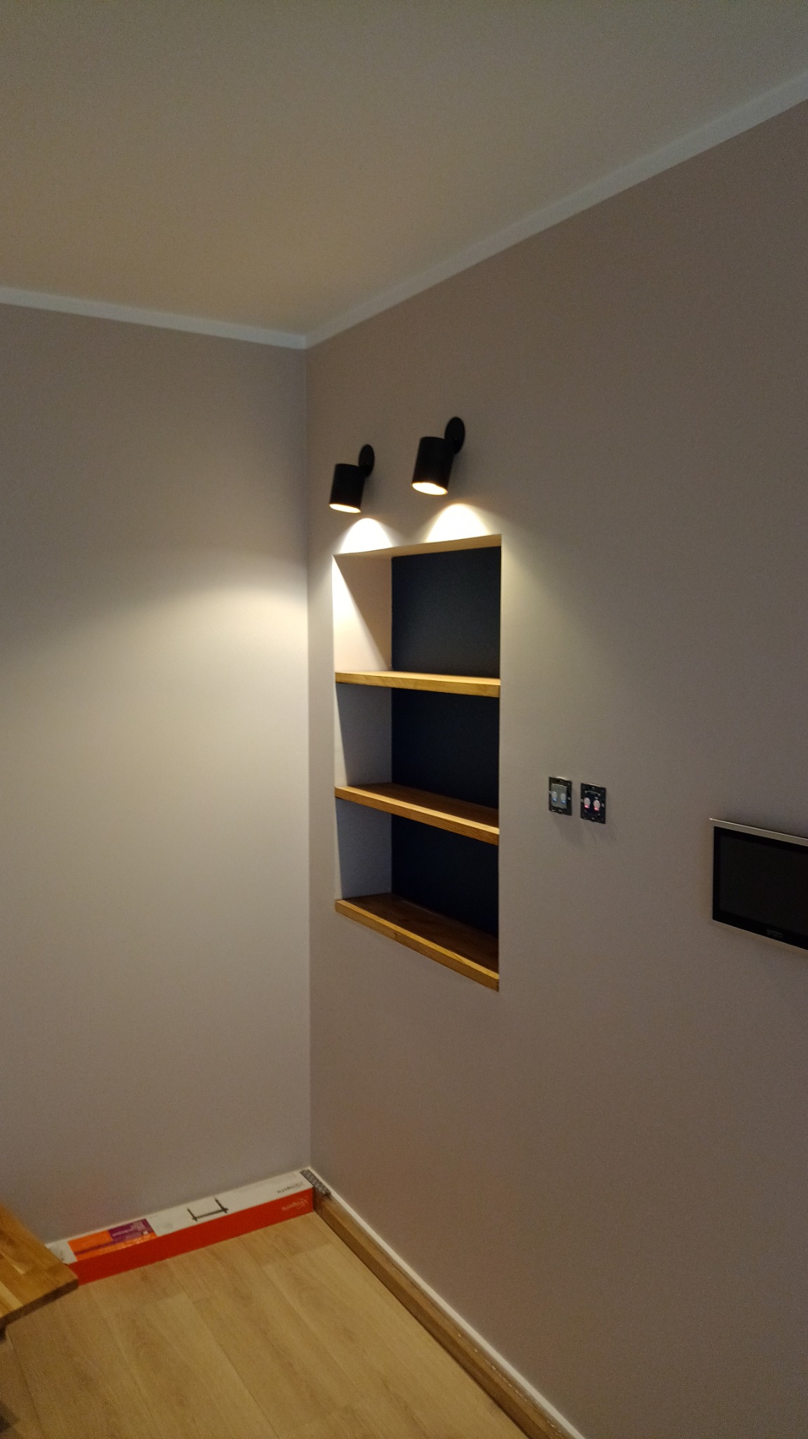 Wnęka ścienna z drewnianymi półkami podświetlona dwoma czarnymi reflektorami punktowymi, ściany w odcieniach szarości, parkiet na podłodze, widoczne gniazdka elektryczne.