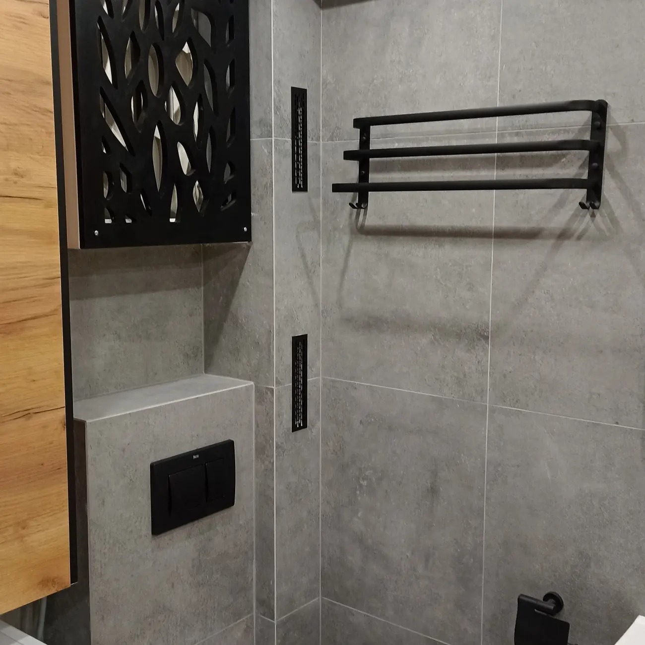 Szare płytki w łazience z czarnymi akcentami: dekoracyjna szafka z ażurowym frontem, grzejnik drabinkowy na ręczniki, przycisk spłuczki podtynkowej i kratki wentylacyjne.