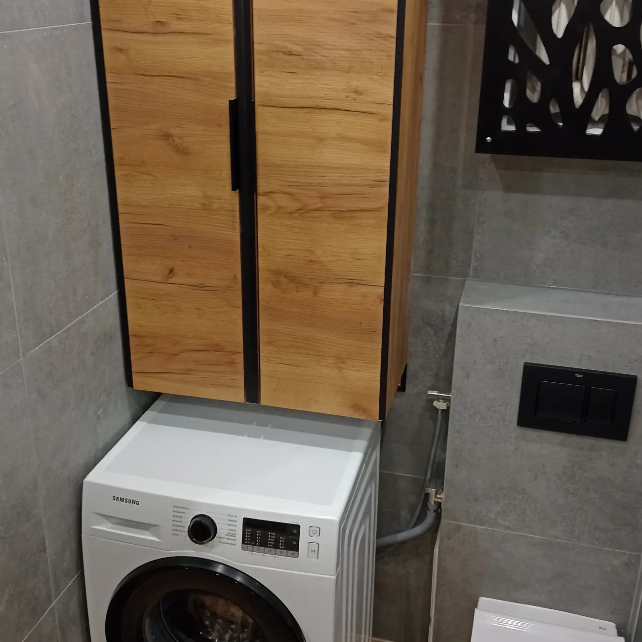 Nowoczesna łazienka z pralką Samsung pod wiszącą szafką z drewnianymi frontami i czarnymi uchwytami, szare płytki na ścianach, czarny panel sterujący spłuczką.