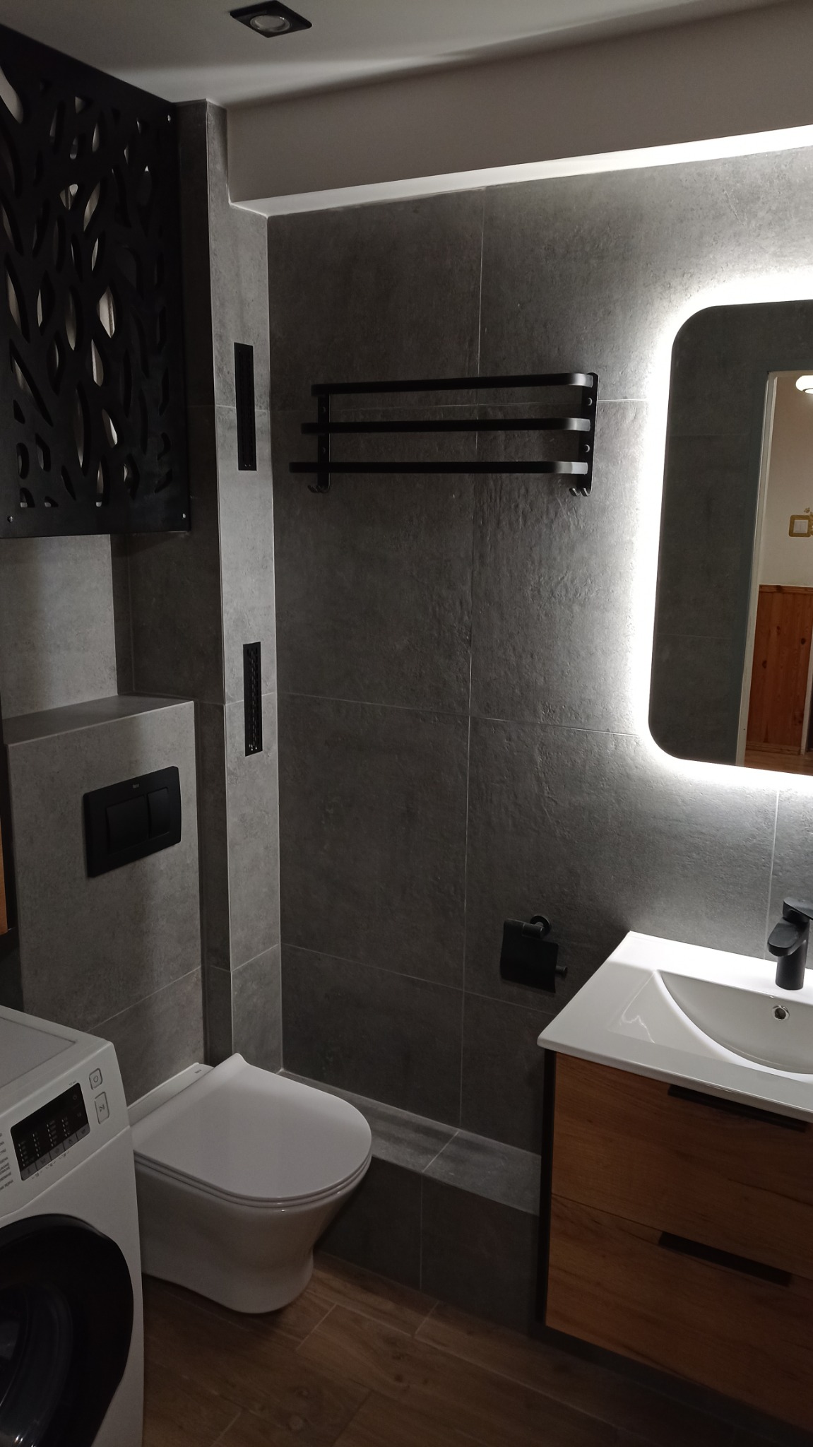 Nowoczesna łazienka z szarymi płytkami, podświetlanym lustrem, drewnianą szafką pod umywalką i dekoracyjnym panelem ażurowym.