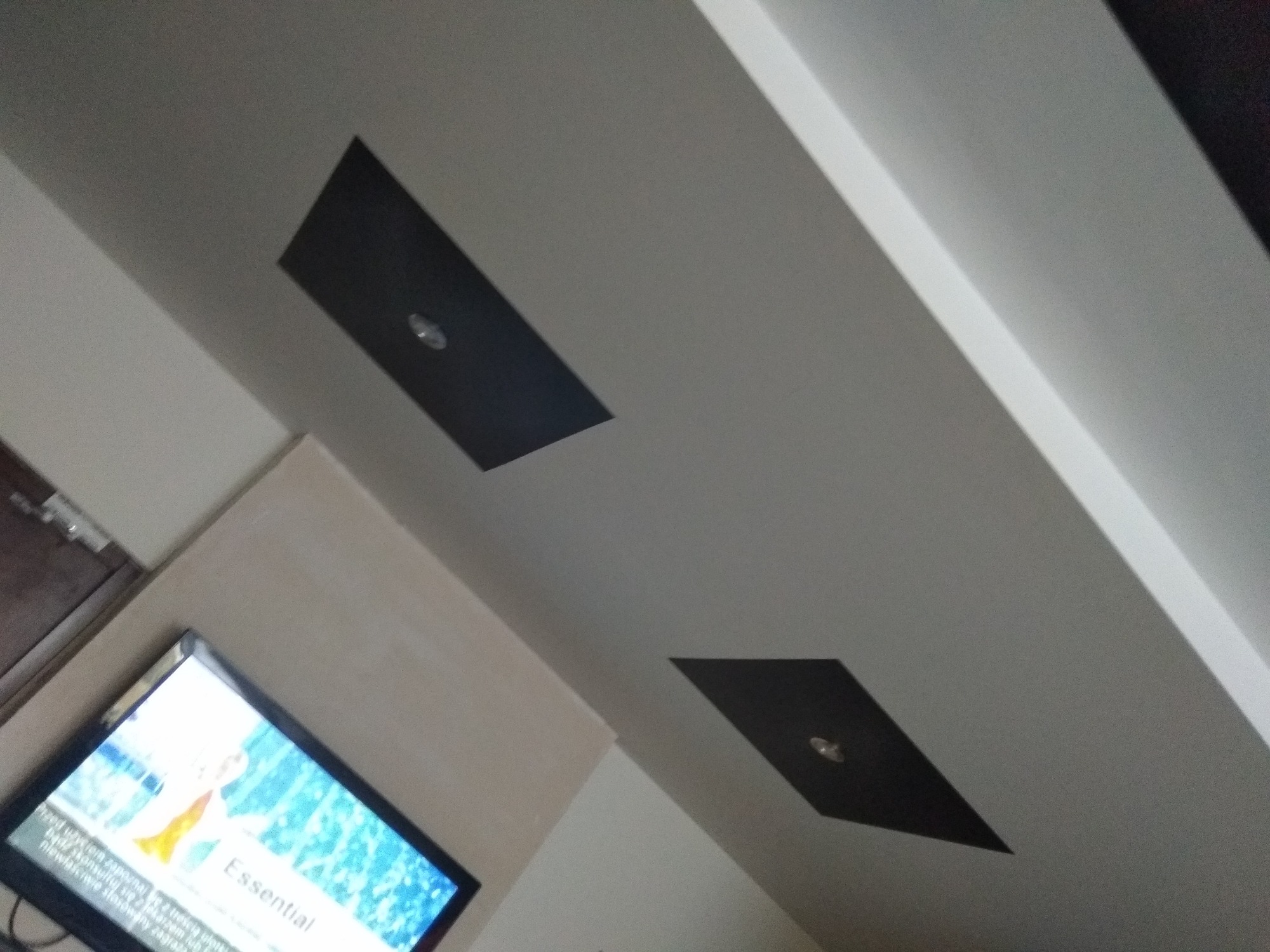 Biały sufit z dwoma prostokątnymi, czarnymi panelami oświetleniowymi z punktowymi lampami, widoczny fragment telewizora w rogu.