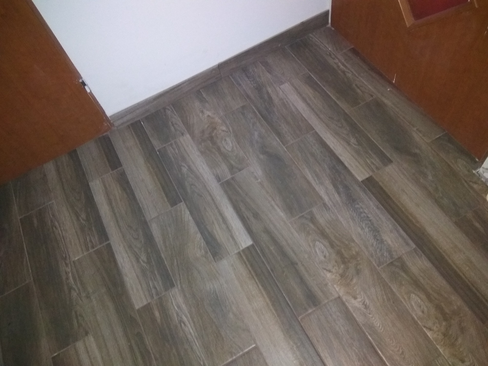Ułożona pod kątem podłoga z płytek imitujących drewno w odcieniach szarości i brązu, widoczna listwa przypodłogowa i fragment brązowych drzwi.