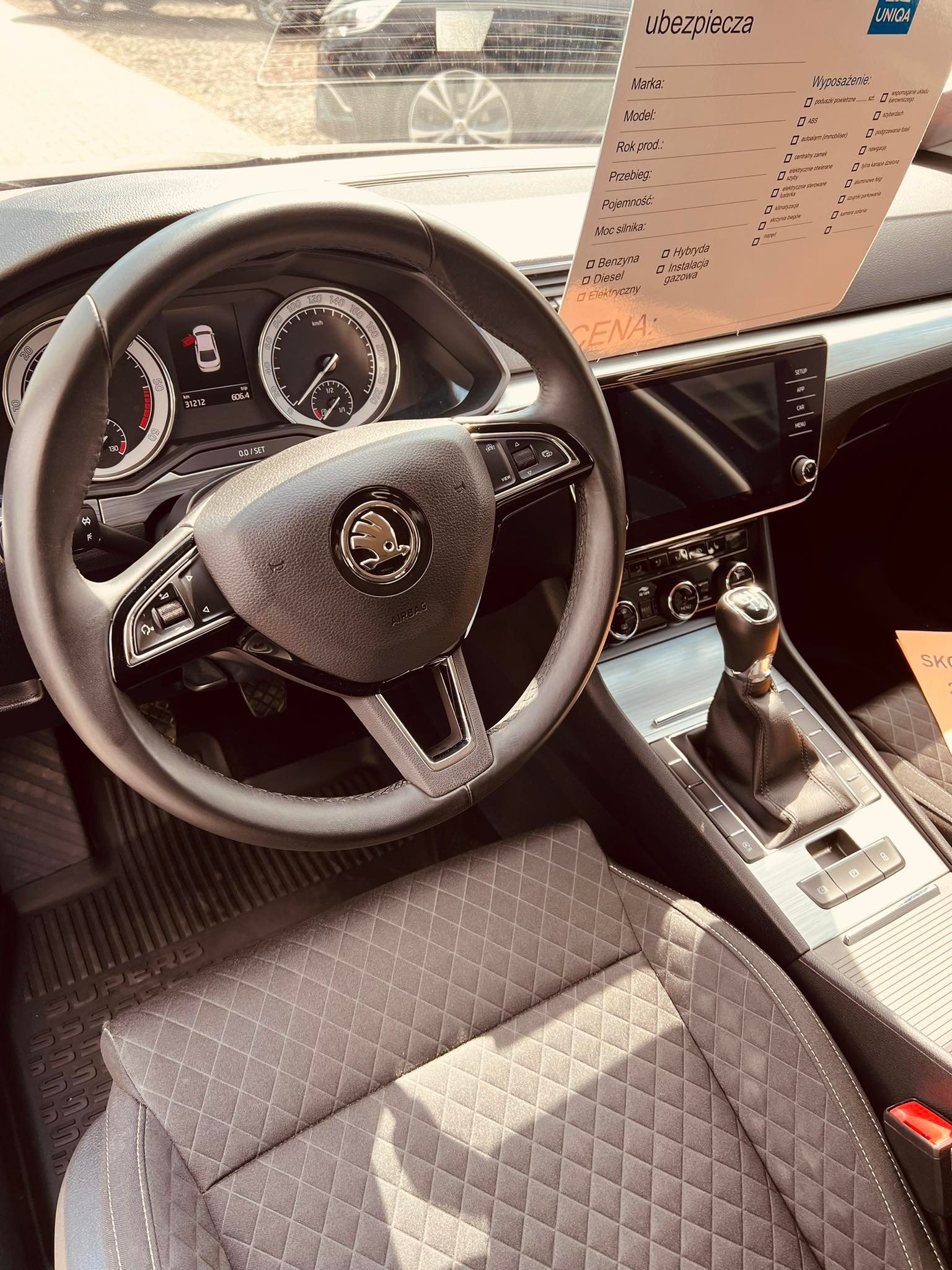 Wnętrze samochodu Skoda Superb z widoczną deską rozdzielczą, kierownicą z logo marki, skrzynią biegów, tapicerką fotela w romby oraz kartką z informacjami o ubezpieczeniu i wyposażeniu pojazdu.