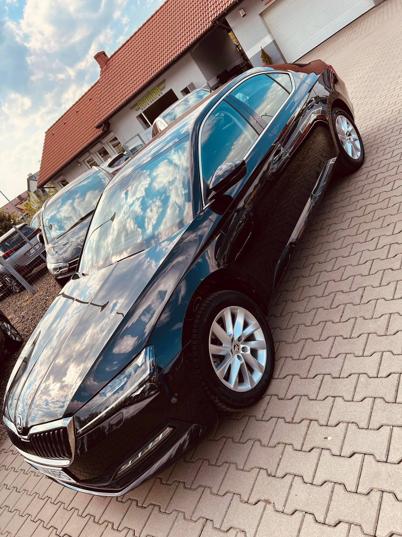 Błyszczący, czarny samochód osobowy marki Skoda Superb, zaparkowany na brukowanym placu przed salonem samochodowym z czerwoną dachówką, odbijający błękitne niebo w karoserii.