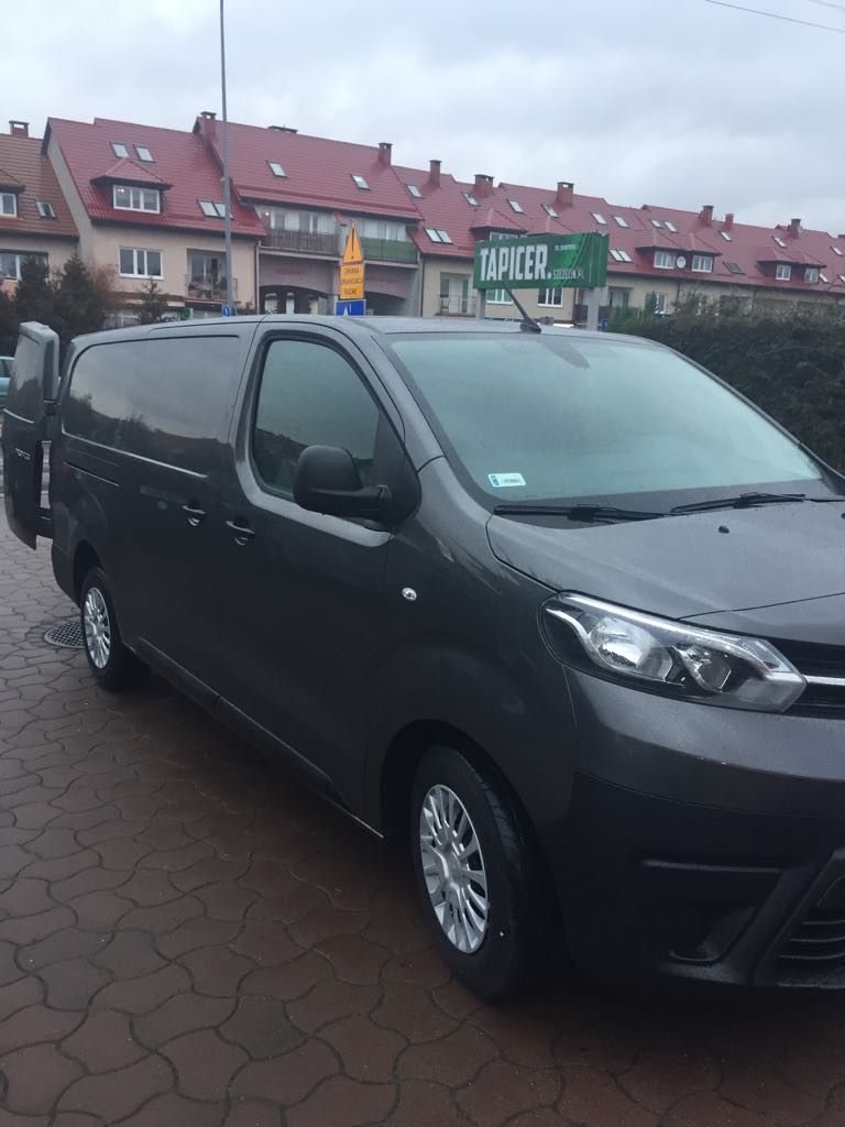 Szary van z otwartymi tylnymi drzwiami na brukowanej ulicy, w tle budynki z czerwonymi dachami i zielony szyld 'TAPICER'.