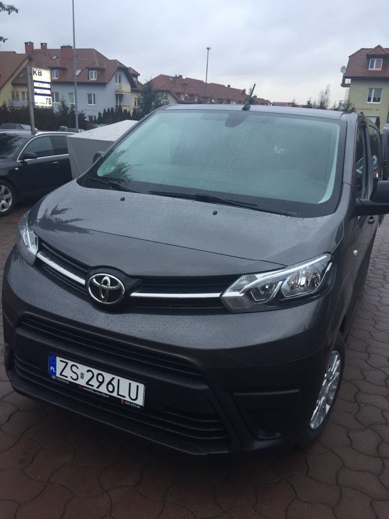 Szary minivan Toyota z polską tablicą rejestracyjną ZS 296LU, zaparkowany na brukowanej nawierzchni w pochmurny dzień, widok z przodu.