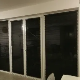 Cztery okna z białymi ramami, zasłonięte czarnymi żaluzjami poziomymi, widoczne w jasnym wnętrzu. Na pierwszym planie fragment sofy.