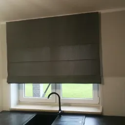 Szara roleta rzymska zamontowana w oknie kuchennym, widok z wnętrza pomieszczenia na zielony trawnik.