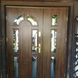 Dwuskrzydłowe, brązowe drzwi wejściowe z przeszkleniami o różnych kształtach, zamontowane w drewnianej ramie.