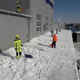 Pracownicy w odblaskowych ubraniach usuwają śnieg z płaskiego dachu budynku przemysłowego za pomocą łopat i odśnieżarki ręcznej, widoczny napis 'B&P Engineering' na elewacji.