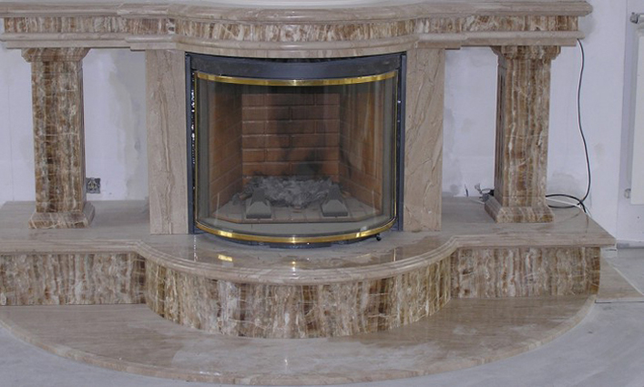 Obudowa kominka wykonana z polerowanego, beżowo-brązowego marmuru z wyraźnym żyłkowaniem, z półokrągłym paleniskiem zabezpieczonym szybą w złotej ramie.