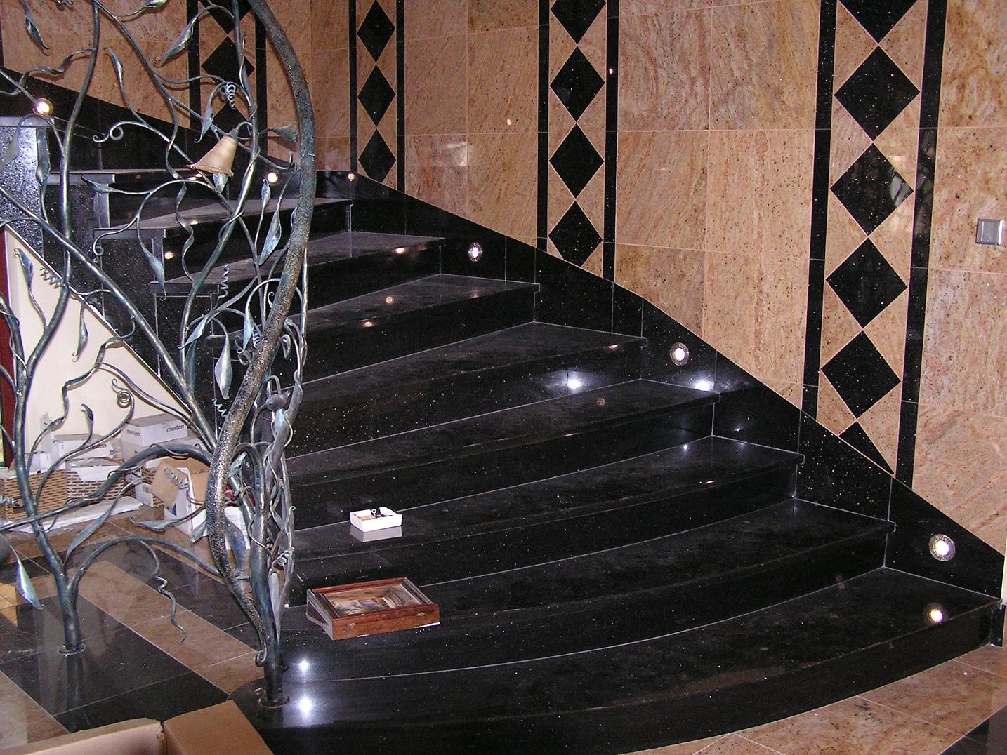 Spiralne schody wyłożone czarnym granitem z dekoracyjną, metalową balustradą w kształcie gałęzi, oświetlone małymi punktowymi lampkami, ściany wykończone płytkami w kolorach beżu i czerni.