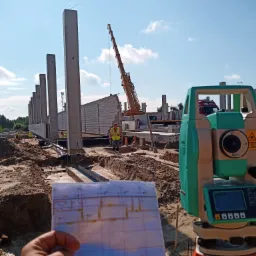 Budowa hali: widok na plac budowy z teodolitem w pierwszym planie i planem w dłoni, w tle dźwig i montowane ściany z prefabrykatów.