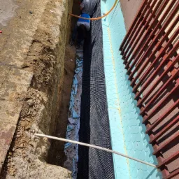 Prace ziemne przy budynku: robotnik w wykopie montuje izolację pionową fundamentów z folii kubełkowej i niebieskiego styropianu, widoczne zbrojenie balkonu.