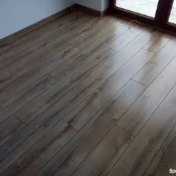 Ułożone panele podłogowe w odcieniach brązu, widok z góry, z widocznym oknem i fragmentem białej ściany.