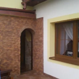 Fragment elewacji budynku z kamienną okładziną wokół łukowego wejścia, brązowe okno z białymi zasłonami, żółte akcenty kolorystyczne, widoczny fragment rynny i płytki na podłodze.