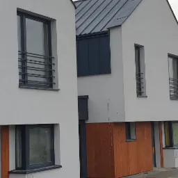 Dwa nowoczesne, białe domy z płaskimi dachami i drewnianymi panelami na elewacji, widoczne czarne ramy okienne i metalowe balustrady okienne, szara, pochmurna pogoda w tle.