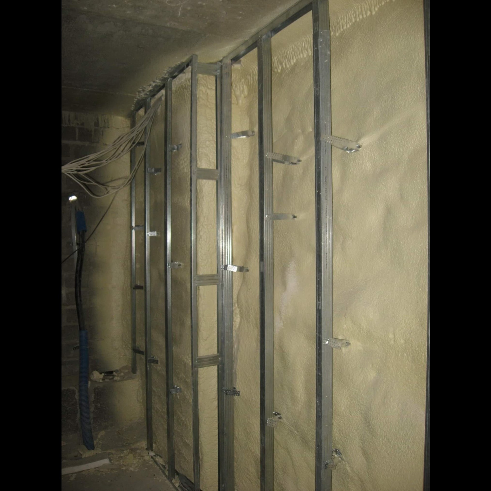 Ściana wewnętrzna z metalowym stelażem i zaaplikowaną warstwą izolacyjnej pianki o porowatej strukturze; widoczne przewody elektryczne w tle.