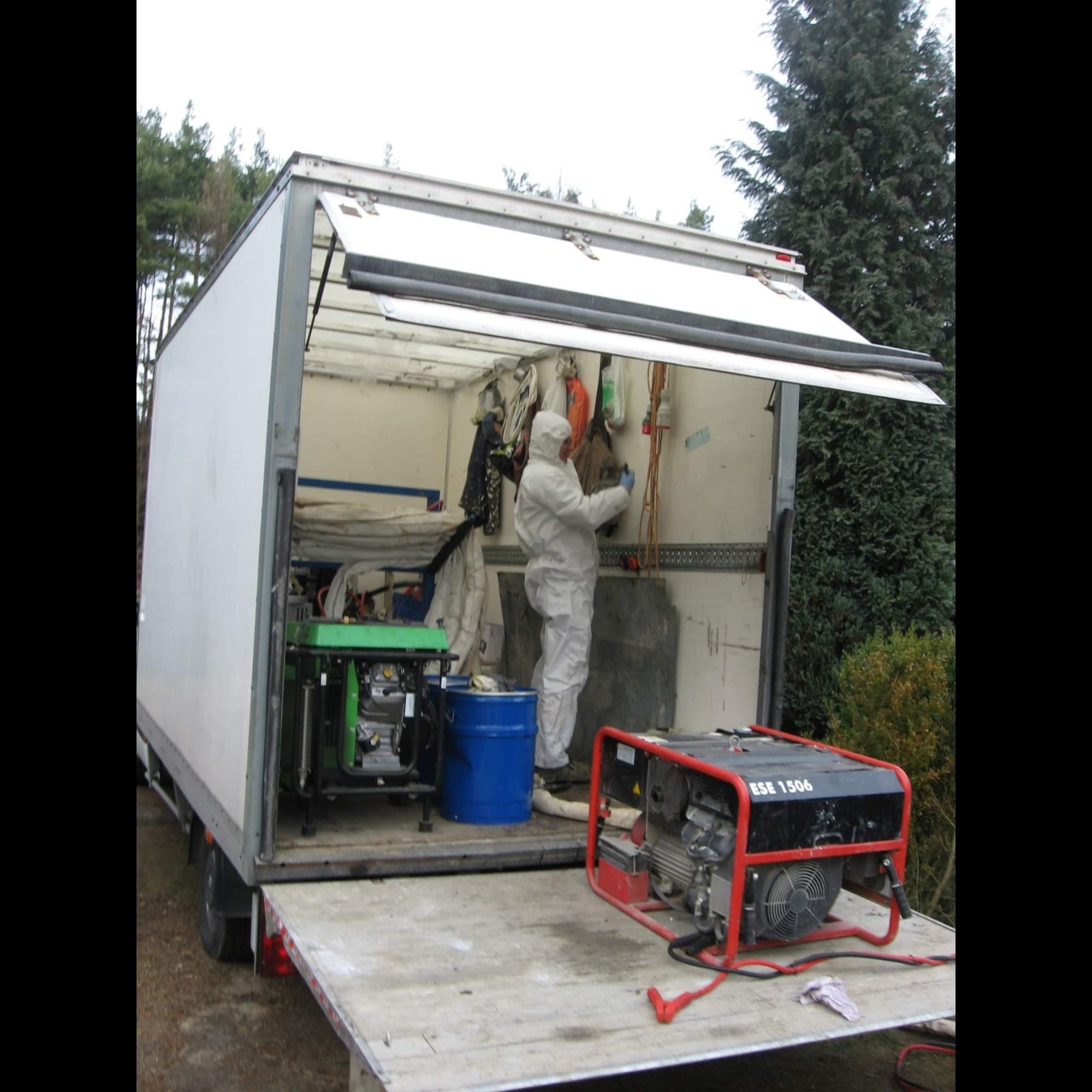 Pracownik w kombinezonie ochronnym przygotowuje się do pracy wewnątrz furgonetki, obok widoczne agregaty prądotwórcze i niebieska beczka.