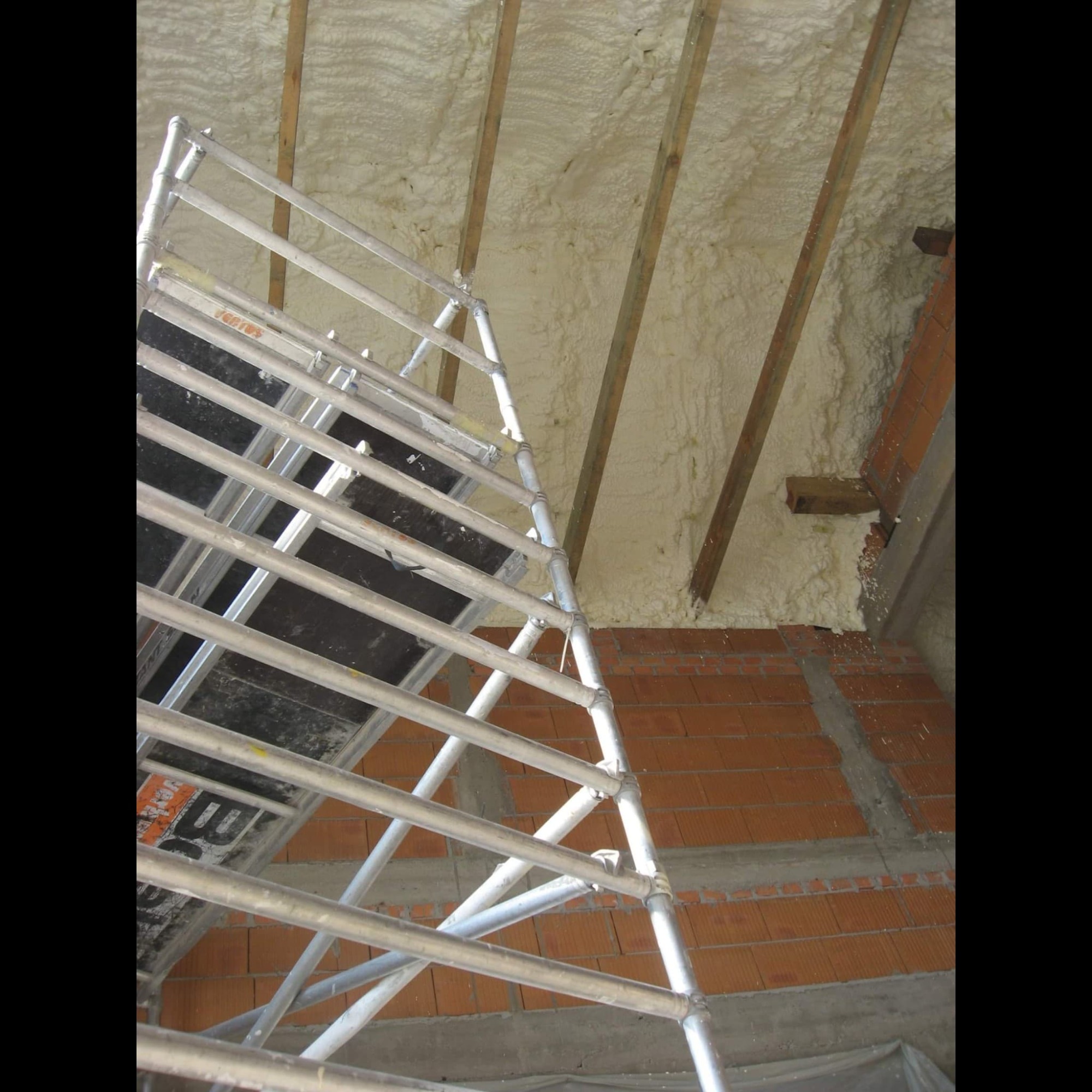 Ocieplanie poddasza pianką PUR widoczne z perspektywy rusztowania, eksponowane drewniane belki stropowe i ceglana ściana.