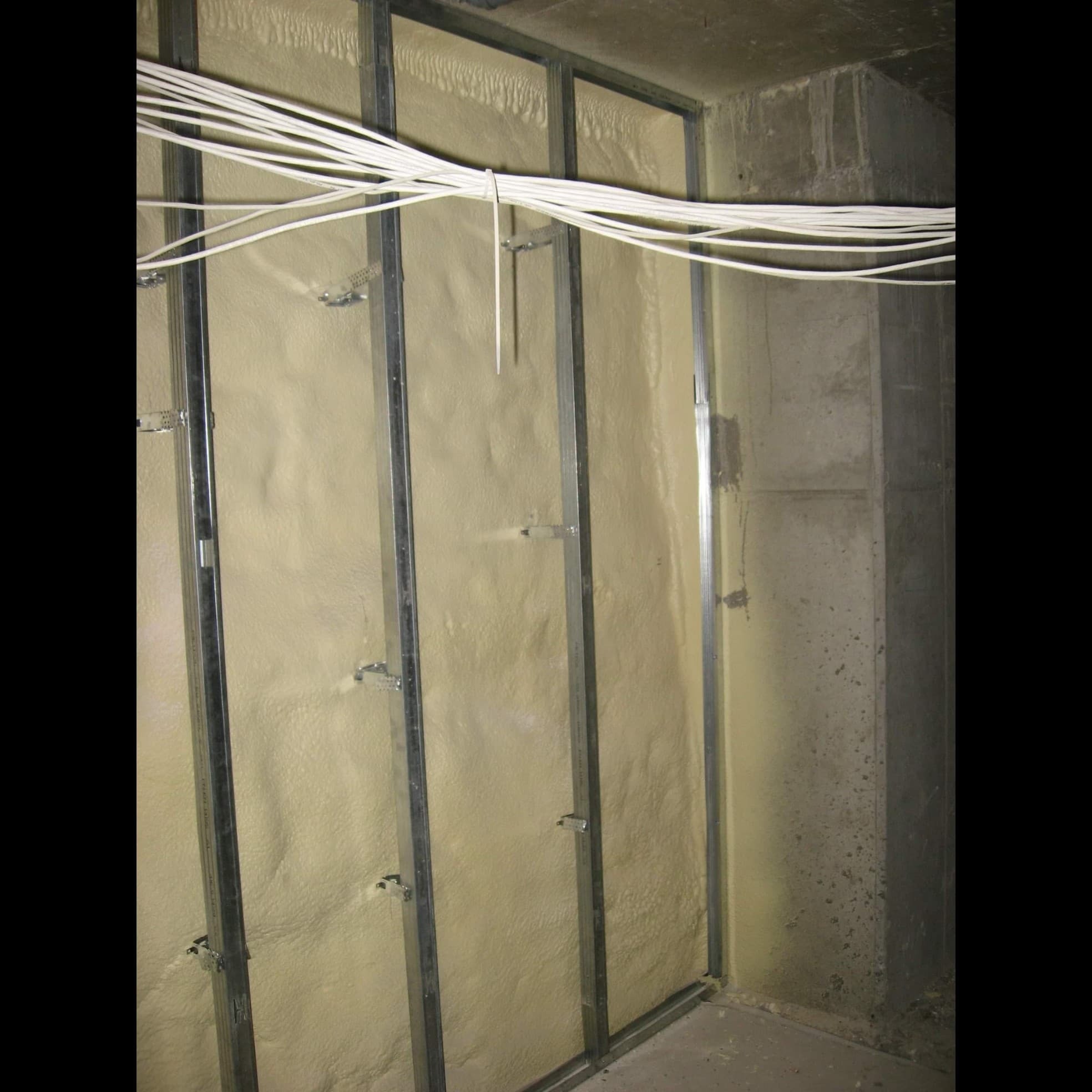Ściana z metalowym stelażem wypełniona pianką izolacyjną, widoczne przewody elektryczne pod sufitem, surowy beton w rogu pomieszczenia.