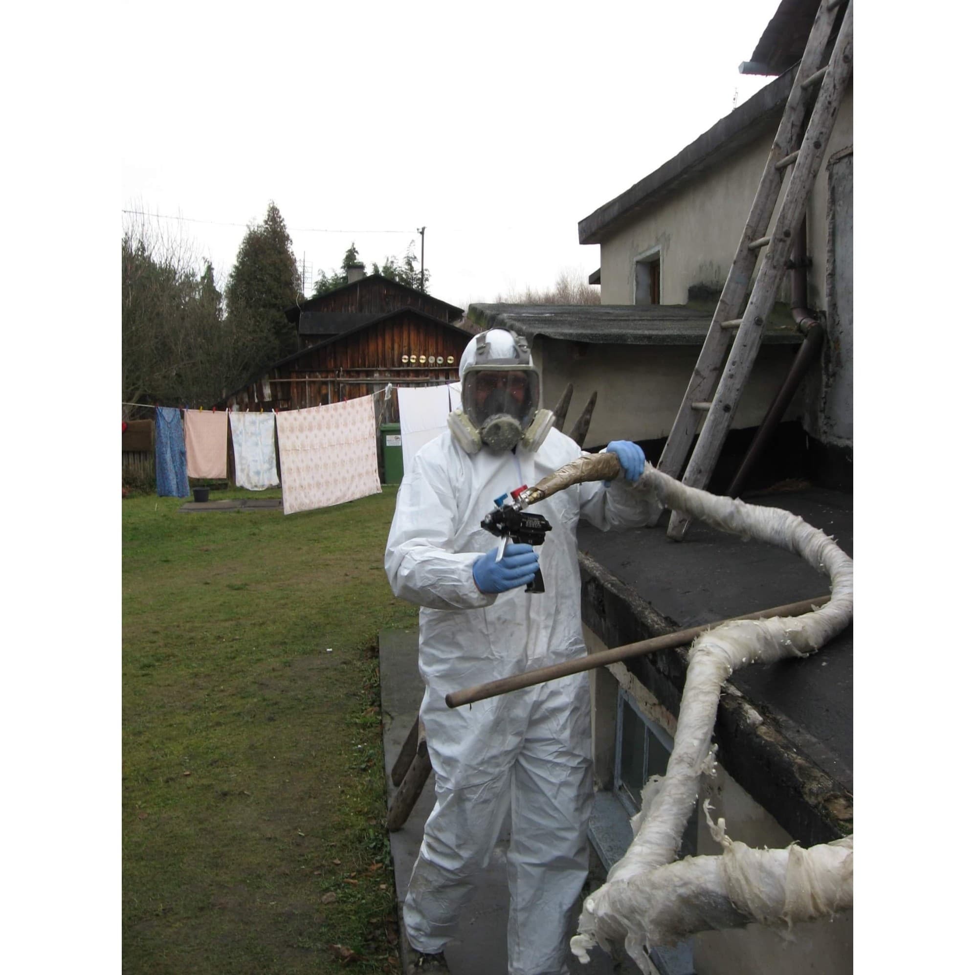 Pracownik w kombinezonie ochronnym i masce, z pistoletem natryskowym, aplikuje izolację na zewnątrz budynku, widoczna drabina oparta o ścianę i pranie suszące się w tle.