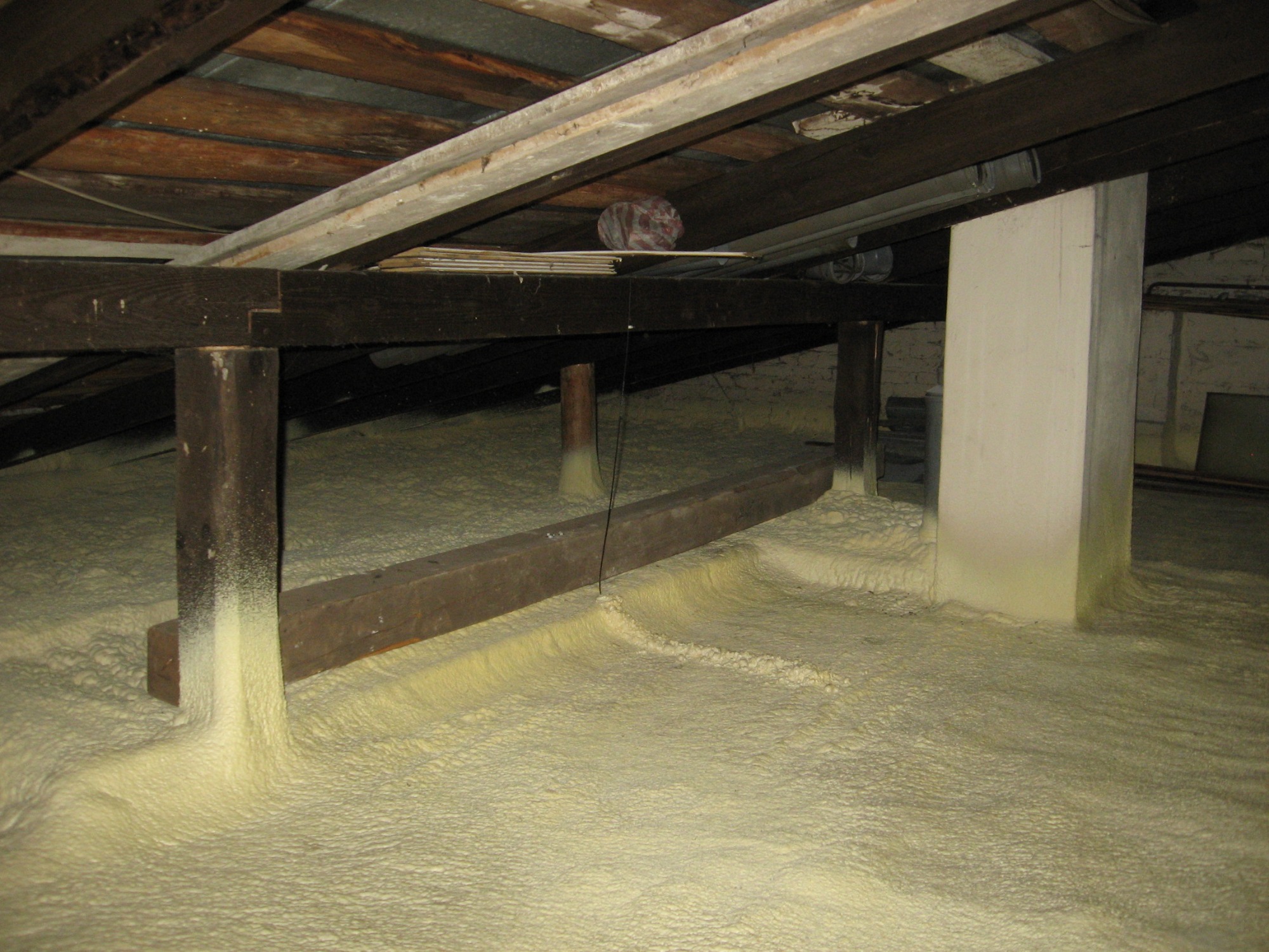 Poddasze z drewnianą konstrukcją pokryte warstwą izolacyjnej pianki natryskowej, widoczne elementy instalacji i oświetlenia, ściany z cegły.