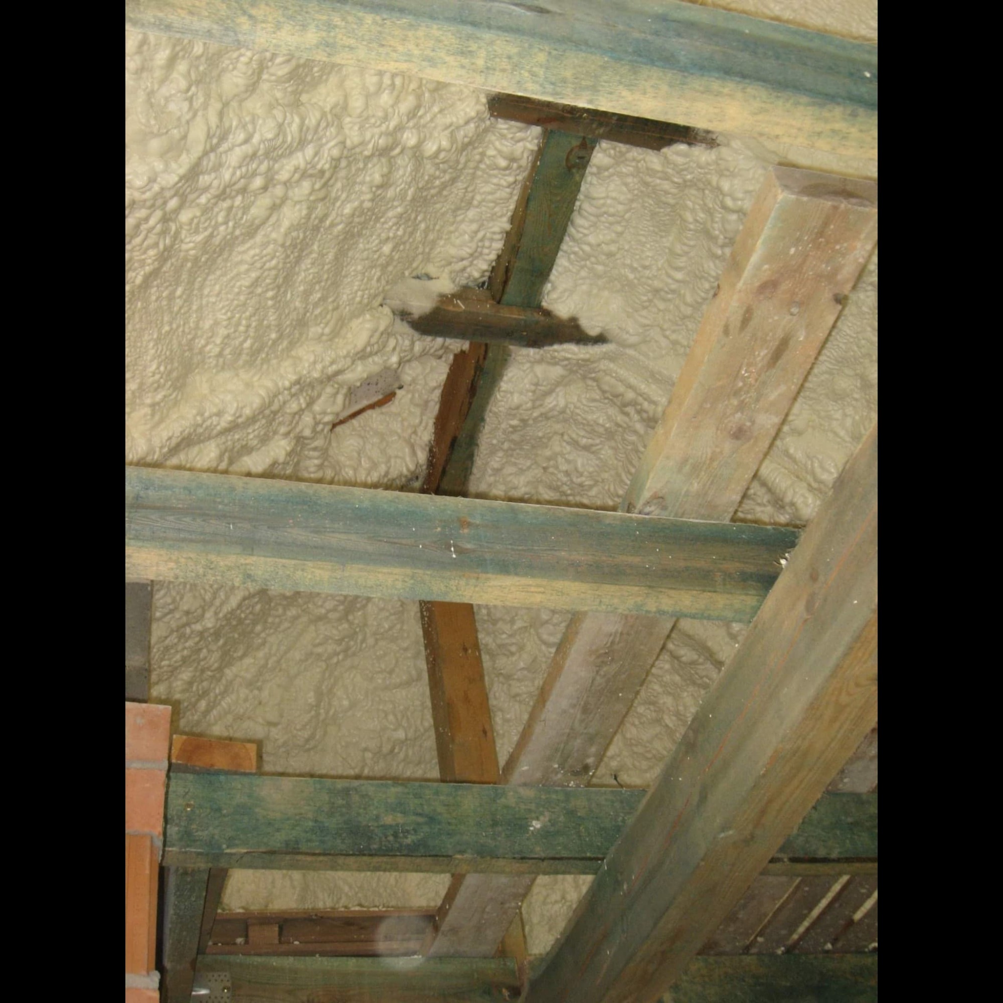 Poddasze ocieplone pianką poliuretanową, widoczne drewniane krokwie i fragment muru z cegły. Niektóre elementy konstrukcji drewnianej są częściowo zakryte pianą.