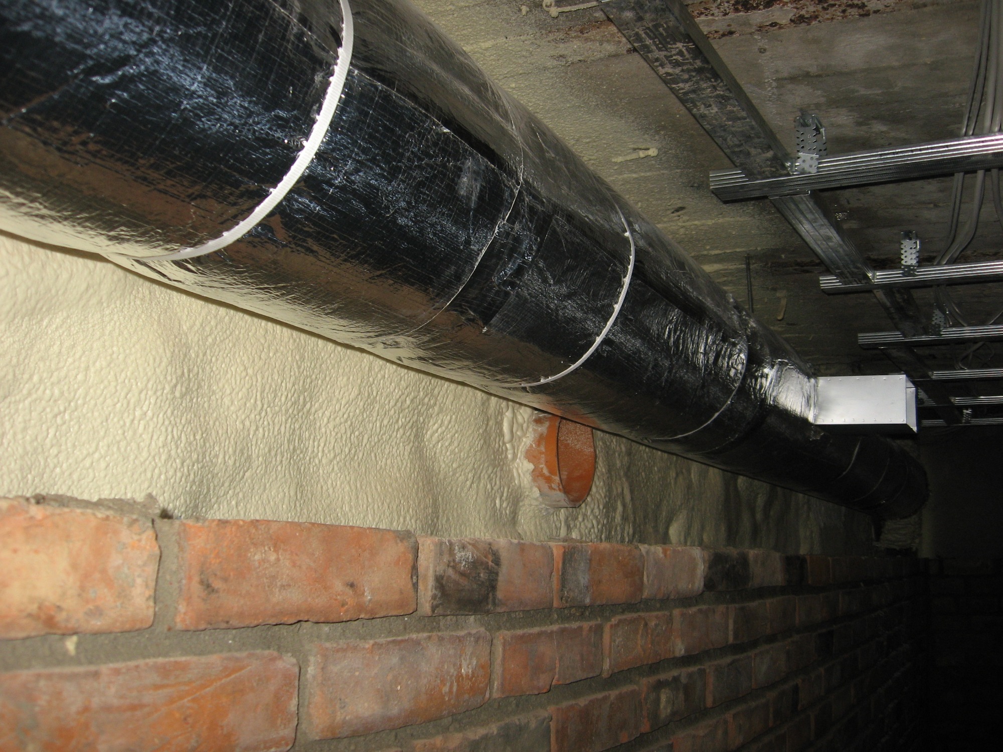 Ocieplony pianką PUR fragment ściany z cegły z widocznym otworem rury i metalową rurą wentylacyjną owiniętą srebrną folią, zamocowaną do sufitu za pomocą metalowych wsporników.