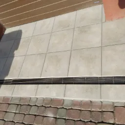 Jasnoszare płytki tarasowe z odpływem liniowym i kostką brukową, widoczne cienie i fragment ściany budynku.