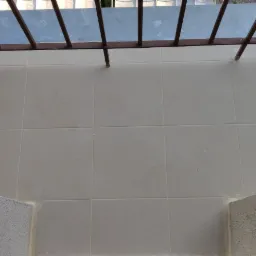 Jasne, kwadratowe płytki ceramiczne na balkonie z szarą balustradą i ścianami o chropowatej fakturze, widoczne ślady ołówka na płytkach.