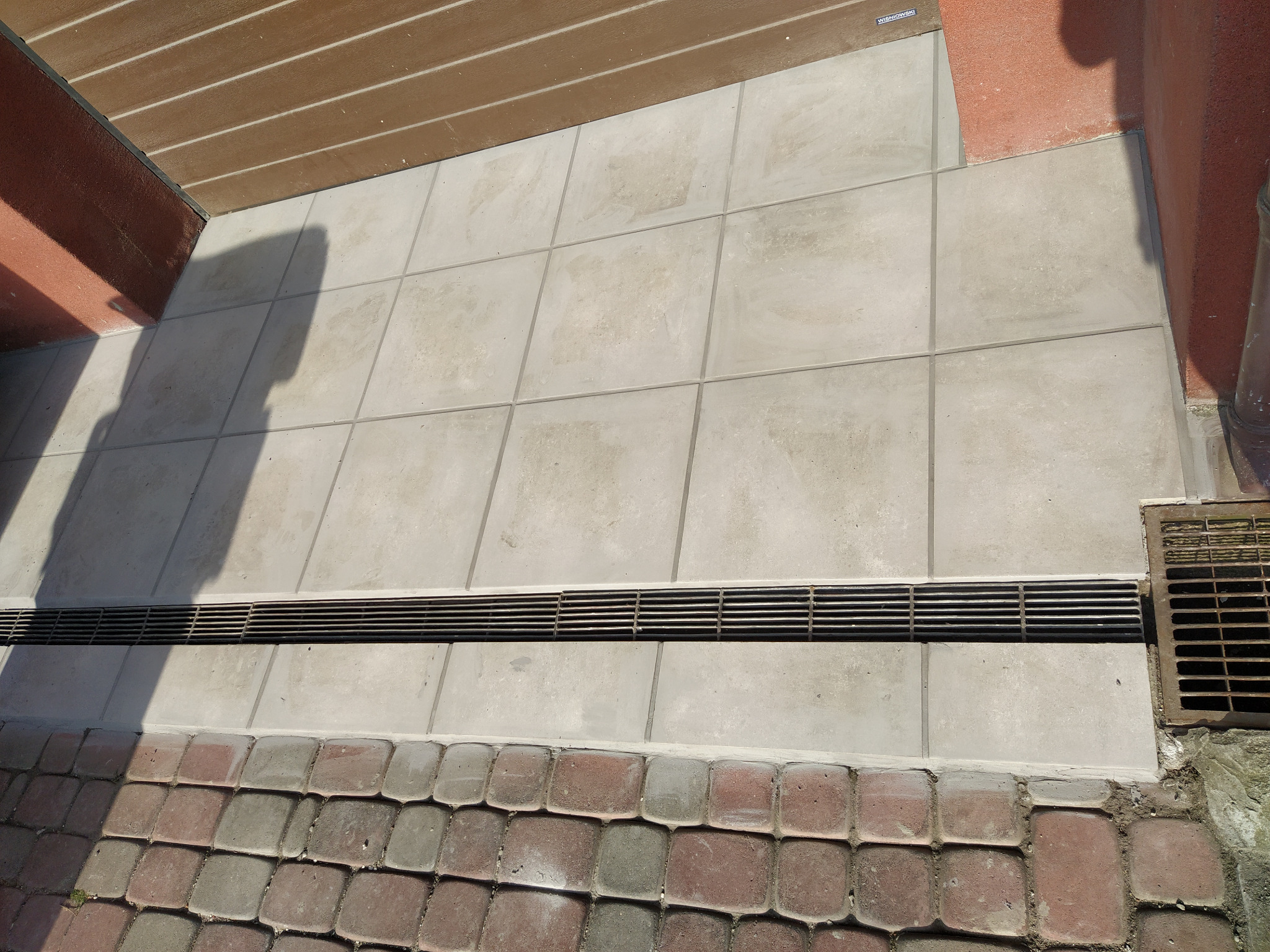 Jasnoszare płytki tarasowe z odpływem liniowym i kostką brukową, widoczne cienie i fragment ściany budynku.