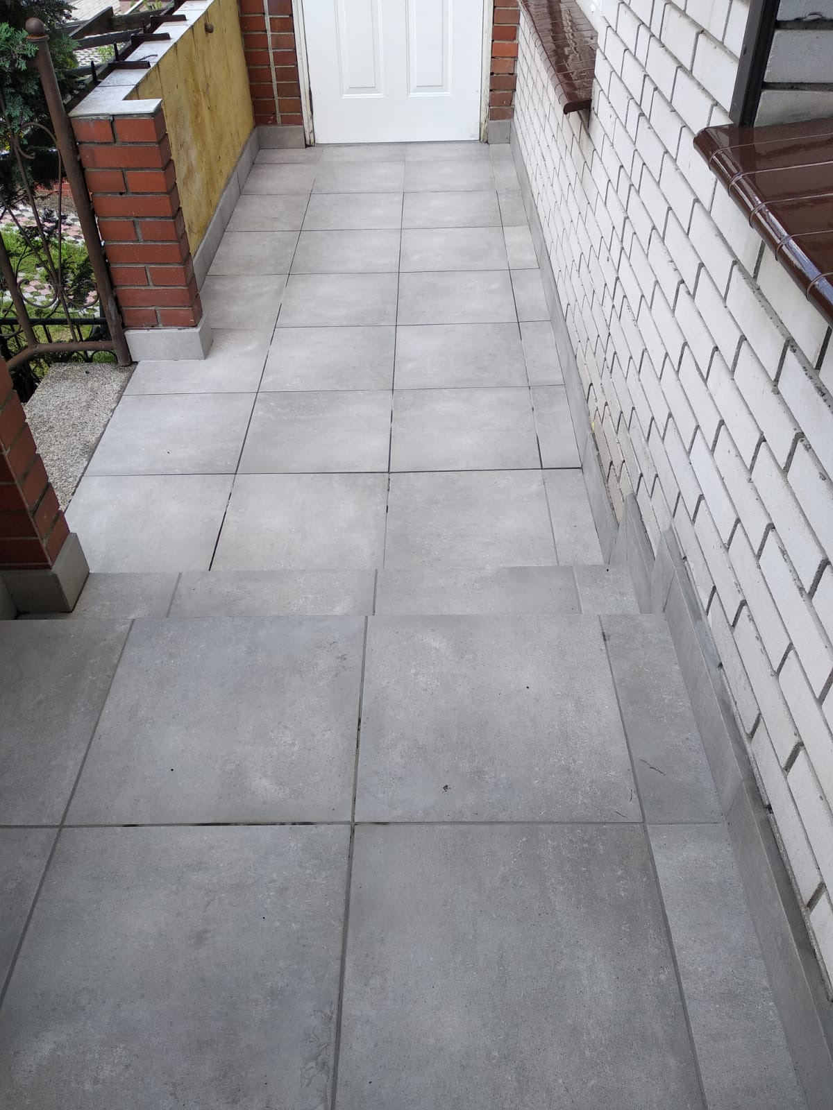 Szare płytki ceramiczne ułożone na schodach wejściowych do domu, z widoczną ścianą z białej cegły i fragmentem balustrady.