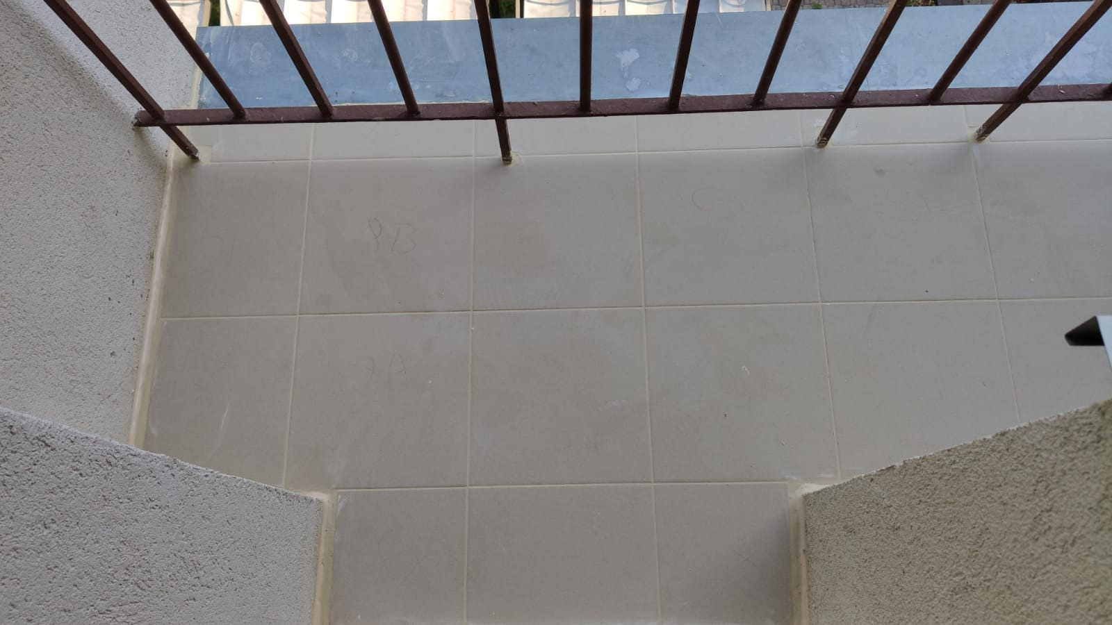 Jasne, kwadratowe płytki ceramiczne na balkonie z szarą balustradą i ścianami o chropowatej fakturze, widoczne ślady ołówka na płytkach.
