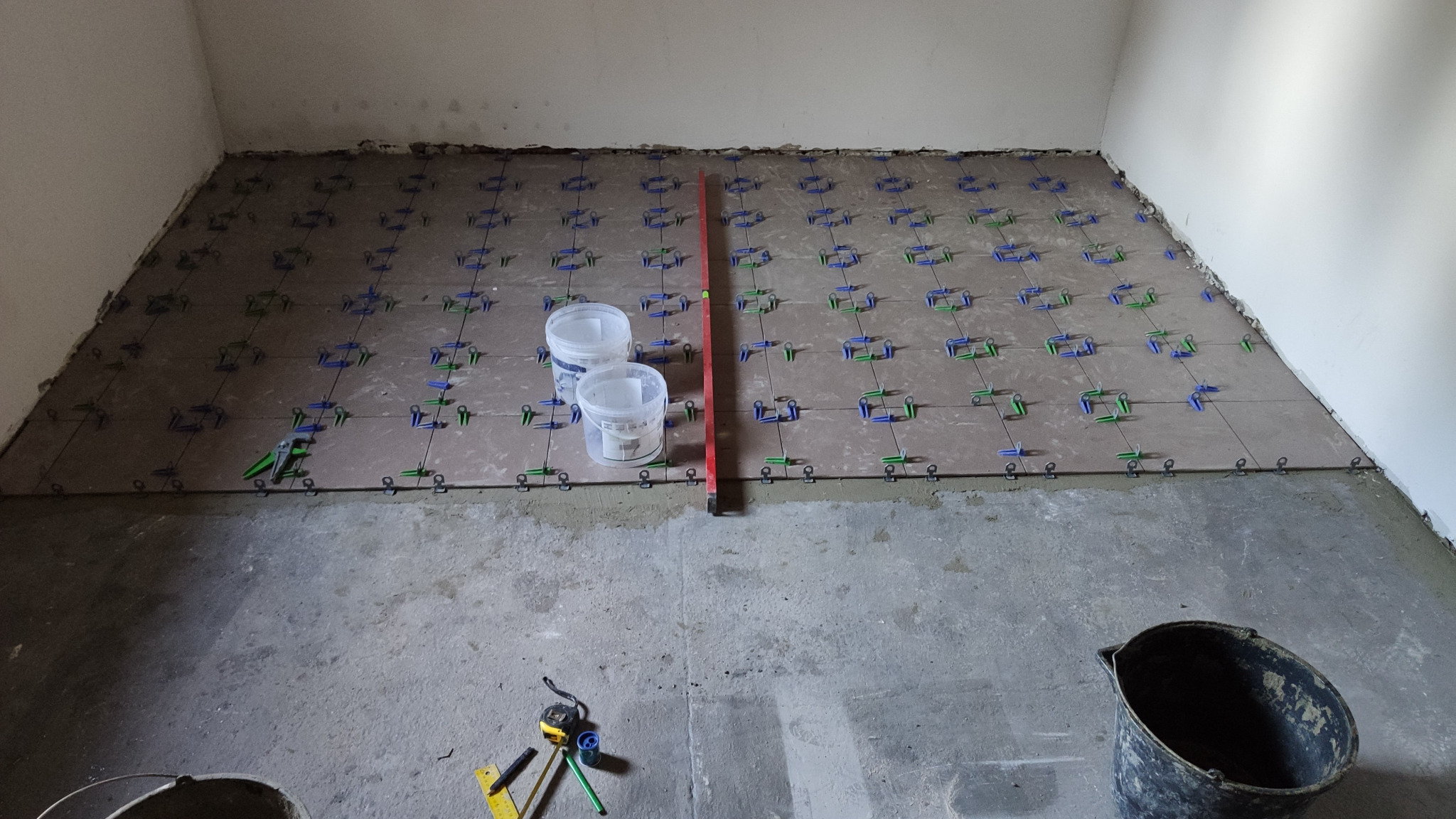 Układanie płytek podłogowych w trakcie prac wykończeniowych, widoczne plastikowe klipsy poziomujące, poziomica, dwa wiadra i narzędzia pomiarowe.