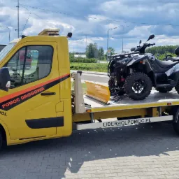 Żółta laweta z napisem 'Pomoc Drogowa Lider Hol' przewozi czarnego quada na platformie, zaparkowana na parkingu z widocznym budynkiem w tle.