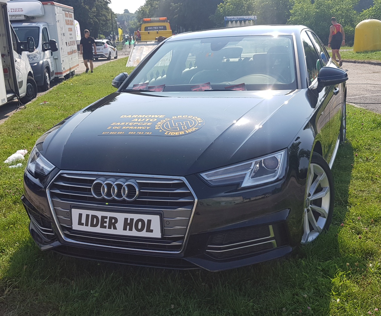Czarny samochód osobowy marki Audi z naklejką reklamową firmy pomocy drogowej na masce i tablicą rejestracyjną 'LIDER HOL' zaparkowany na trawie, w tle pojazdy serwisowe i osoby.