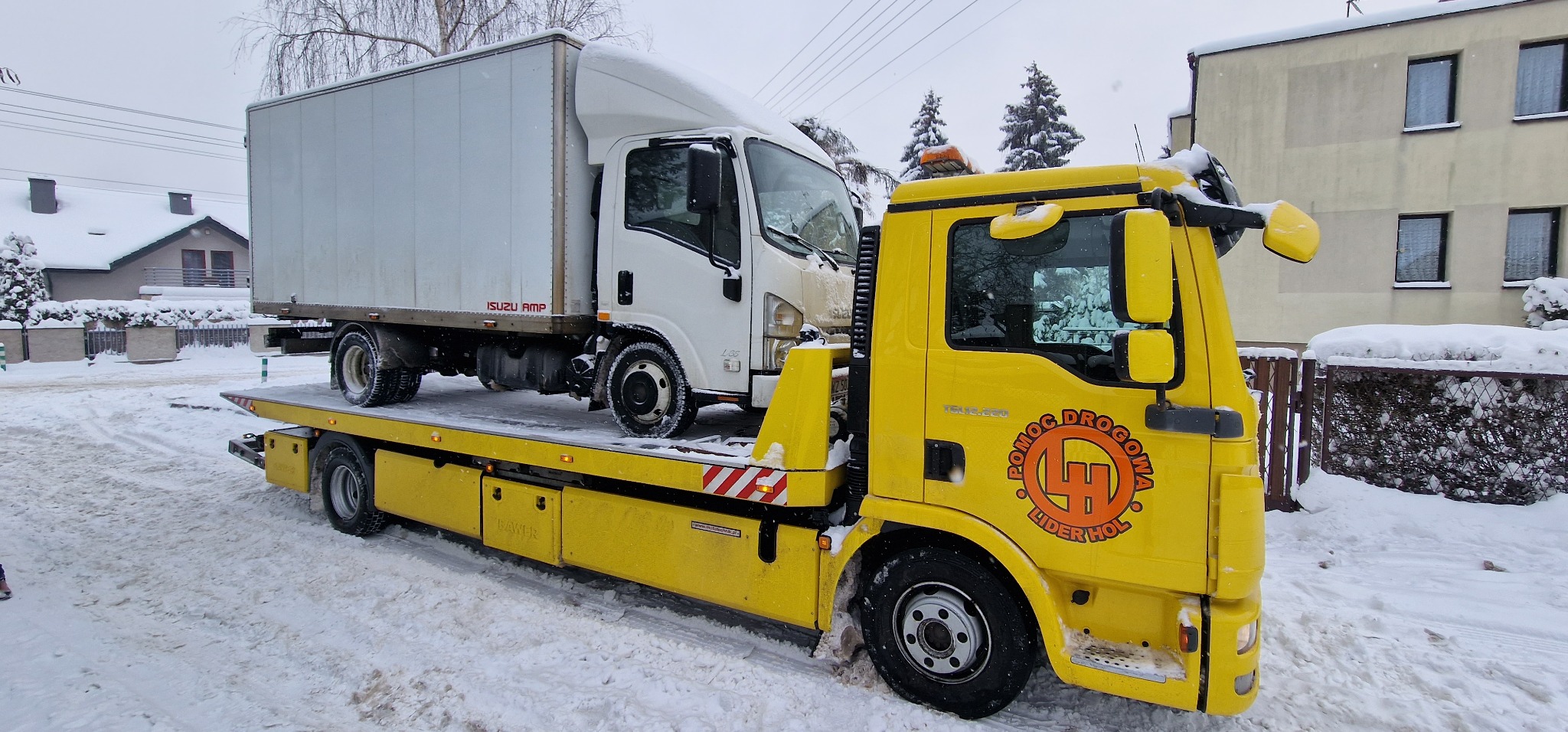 Żółta laweta z logo 'Pomoc Drogowa Lider Hol' przewozi biały samochód dostawczy Isuzu na zaśnieżonej ulicy.