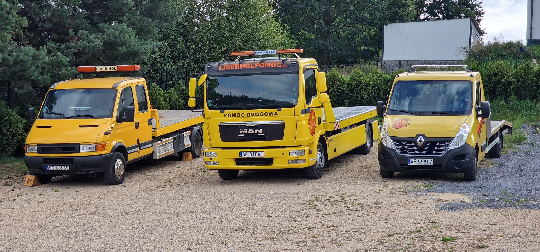 Trzy żółte lawety różnych marek (Iveco, MAN, Renault) zaparkowane na żwirowym placu, z widocznymi tablicami rejestracyjnymi i logotypami firm świadczących pomoc drogową.