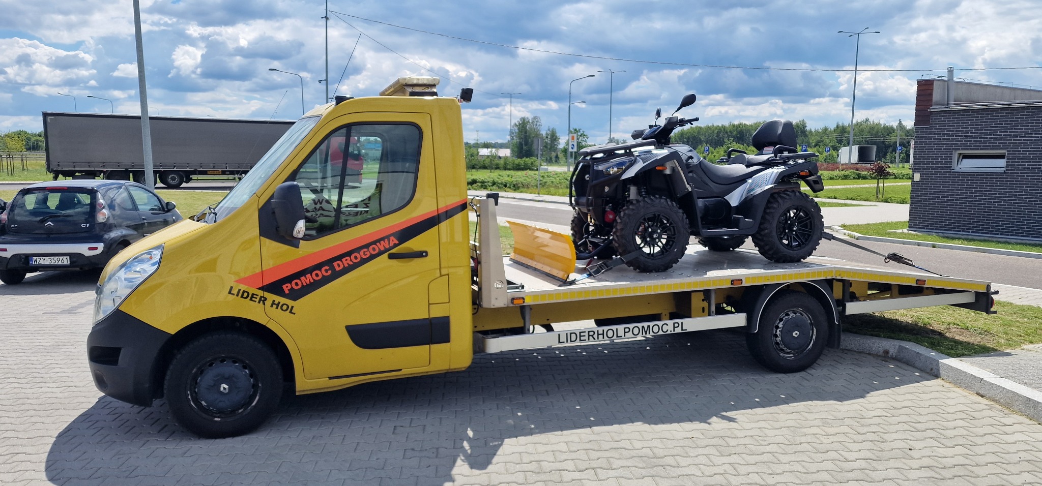 Żółta laweta z napisem 'Pomoc Drogowa Lider Hol' przewozi czarnego quada na platformie, zaparkowana na parkingu z widocznym budynkiem w tle.