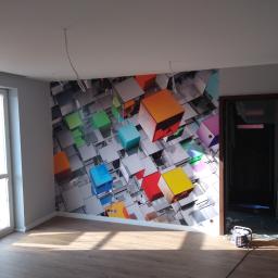 KABREM USŁUGI REMONTOWO BUDOWLANE KONRAD PAWLIK - Pokój po remoncie z panelami podłogowymi i fototapetą z motywem kolorowych kostek, widoczne wiszące przewody elektryczne i otwarte drzwi do kolejnego pomieszczenia.