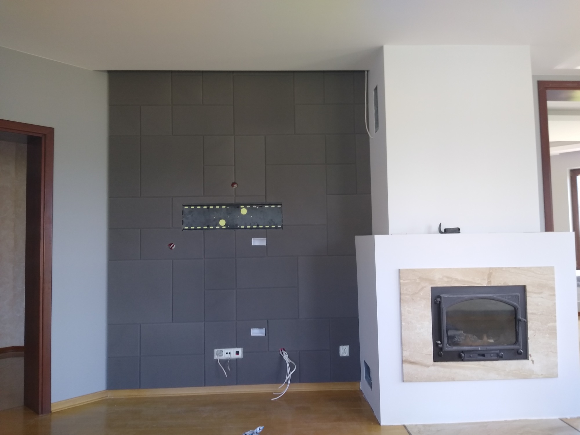 Ściana z szarych paneli z widocznymi puszkami elektrycznymi i przewodami, przygotowana pod montaż telewizora i kominek z marmurową obudową.