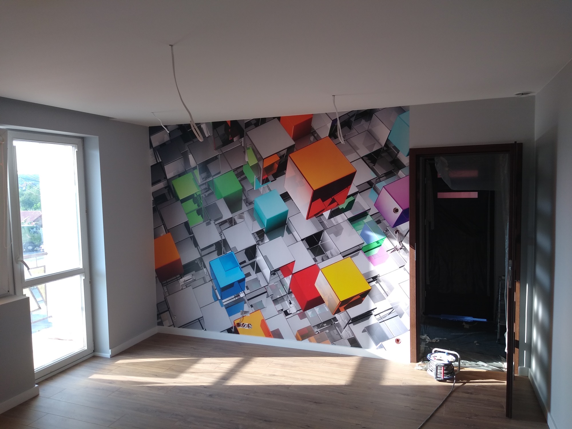 Pokój po remoncie z panelami podłogowymi i fototapetą z motywem kolorowych kostek, widoczne wiszące przewody elektryczne i otwarte drzwi do kolejnego pomieszczenia.