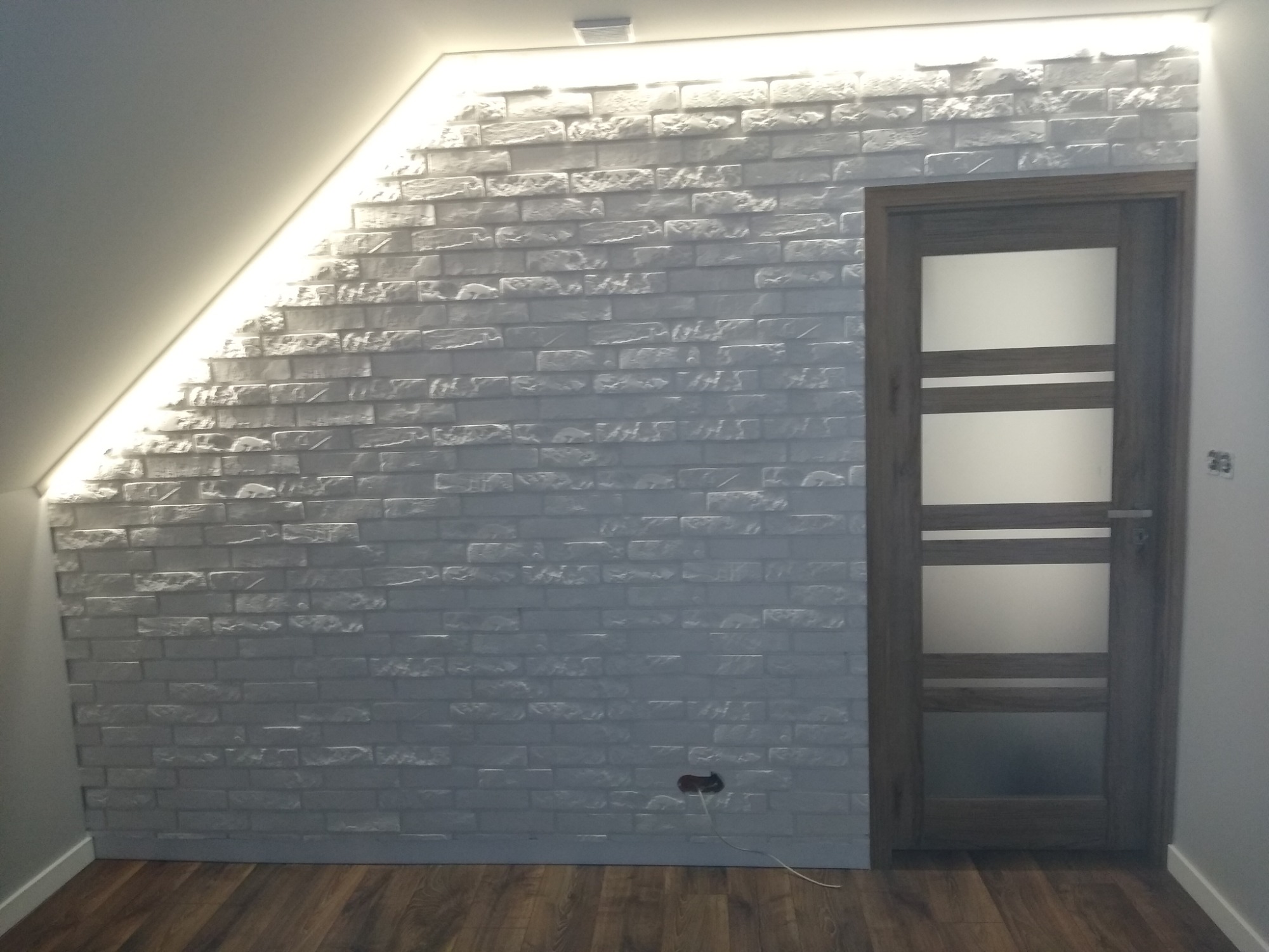 Ściana z białej cegły dekoracyjnej pod skosem, oświetlona od góry ledowym paskiem, obok drewniane drzwi z mlecznym szkłem i brązowe panele podłogowe.